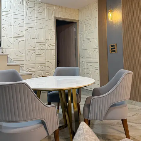 Dining nook with a round marble-top table on brass legs, grey channel-tufted velvet chairs, and a 3D geometric-textured wall panel beside a staircase.