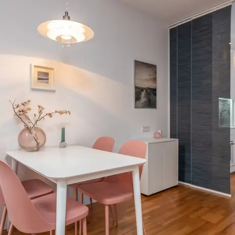Dining nook with a white table and blush-pink chairs beneath a statement pendant light, with framed art and a sliding fabric panel dividing the kitchen beyond.