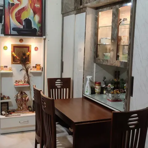 Dining nook with a wood table and chairs beside a marble-laminate crockery unit with profile lighting, adjoining a fitted pooja shelf.
