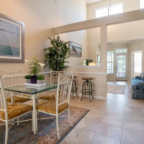 Dining nook with a wrought-iron glass-top table and floral-cushioned chairs on a patterned rug, opening through a half-wall pass into a living room beyond.