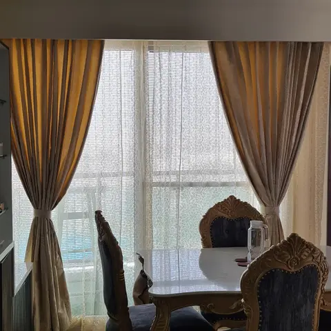 Dining nook with carved gold-finish chairs upholstered in dark navy velvet, a marble-top table and layered gold and beige floor-length curtains at the window.