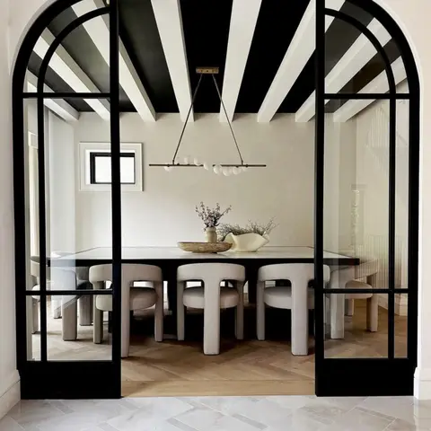 Dining room seen through arched black-framed French doors, with a black-and-white painted exposed-rafter ceiling and a globe-bar chandelier over upholstered