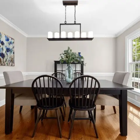 Dining room with a black wood table and spindle-back chairs beneath a linear candle-style chandelier, a black sideboard, and a large blue floral painting.