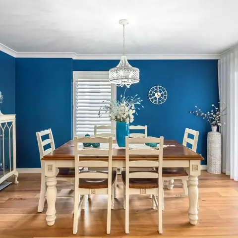 Dining room with a bold blue accent wall, a cream farmhouse-style table and ladder-back chairs, and a woven pendant chandelier.