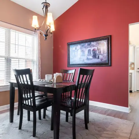 Dining room with a bold red accent wall hung with a framed street-scene painting, a black wood table and slat-back chairs on a patterned grey rug beneath a