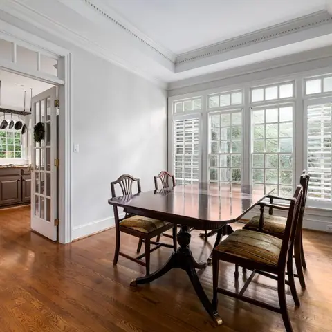 Dining room with a dark wood pedestal table and striped-cushion chairs, tall plantation-shuttered windows, and a French door leading to the kitchen.
