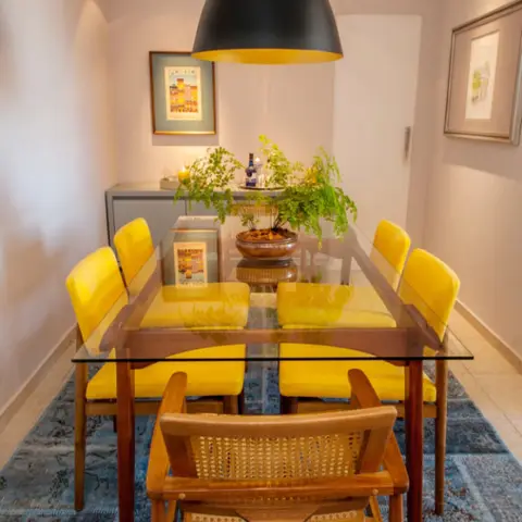 Dining room with a glass-top table, mustard-yellow upholstered chairs, a cane-back armchair and a black dome pendant light
