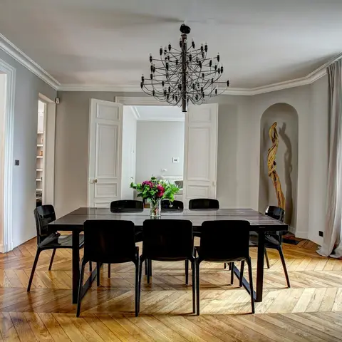 Dining room with a herringbone parquet floor, ornate white panelled doors and a black dining set beneath a statement sputnik chandelier.