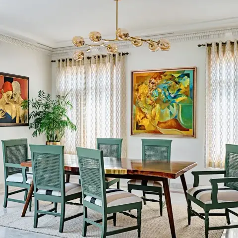 Dining room with a live-edge wood table, sage-green cane-back chairs, a branching gold pendant chandelier, and two large cubist paintings on the wall.