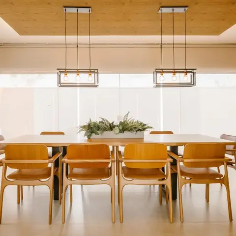 Dining room with a long wood table, eight bentwood armchairs, twin cage pendant lights and a wood-clad ceiling coffer, exposed brick wall visible beyond.