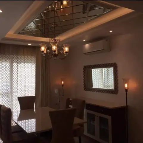 Dining room with a mirror-panelled ceiling insert, crystal chandelier, ornate framed wall mirror, and patterned sheer curtains.