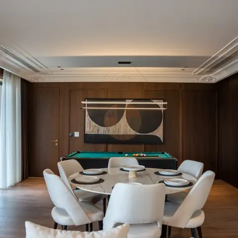 Dining room with a round marble table and white upholstered chairs set for a meal, framed by dark wood panelled walls, a coffered cove-lit ceiling