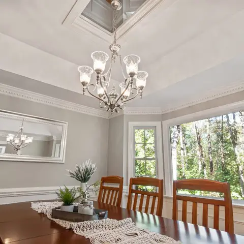 Dining room with a tray-ceiling chandelier, wainscoted grey walls, and a dark wood table beside large windows overlooking wooded grounds.