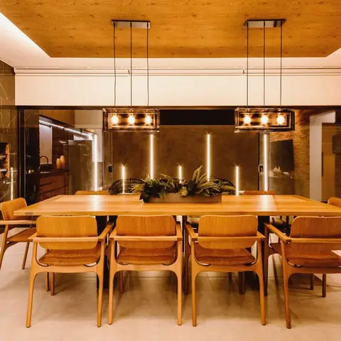 Dining room with a warm wood-plank ceiling, twin box pendant lights over a long wood table with eight curved-arm chairs, and a brick accent wall to the side.