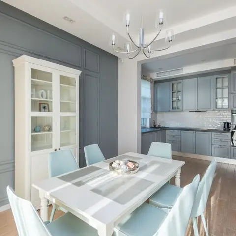 Dining room with a white extendable table and mint-blue chairs against a panelled grey wall, opening through to a blue-grey kitchen.
