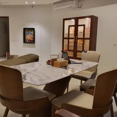 Dining room with a white marble-top table and wood chairs beside a cane-and-glass display cabinet holding pooja idols, with a Tanjore painting on the wall.
