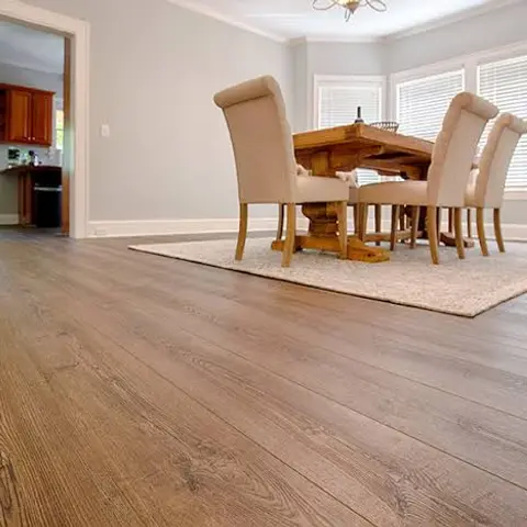Dining room with a wide-plank wood floor, a wood trestle table with upholstered chairs on a pale rug, and a kitchen doorway visible beyond