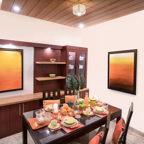 Dining room with a wood-panelled ceiling, a mirror-image pair of sunset-toned framed prints, and a built-in crockery unit beside a styled dining table.