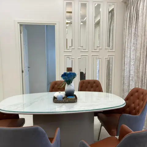 Dining room with an oval marble-top table, tufted brown and grey chairs, a mirrored geometric panel wall, and floor-to-ceiling textured curtains.