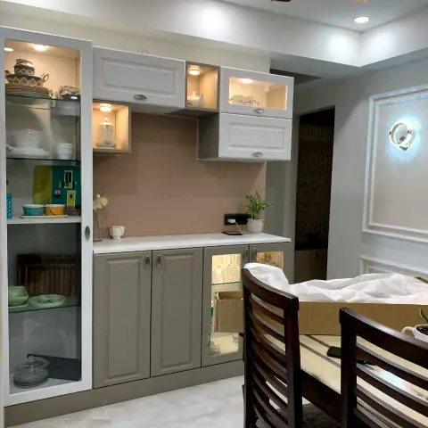 Dining-side crockery unit in greige shaker shutters with a lit glass display tower of dinnerware and a blush back panel over the counter
