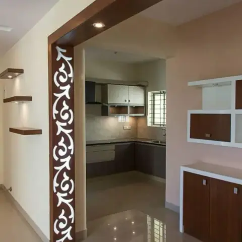 Dining side of a flat looking through a walnut-framed arch with a white CNC jaali strip down one jamb, into the L-shaped kitchen