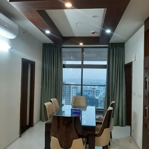 Dining space with a six-seater glass-top table, a wood-boxed false ceiling frame, and floor-to-ceiling windows framed by sage-green curtains overlooking the
