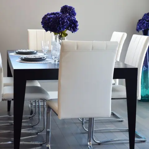 Dining table set with white leather cantilever chairs, a dark wood table with a purple hydrangea centerpiece, and a teal glass vase in the corner