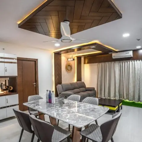 Dining table under a large herringbone wood ceiling raft edged in cove light, with a matching herringbone panel behind the recliner sofa in the living zone