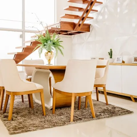 Dining table with cream upholstered chairs on a shag rug beneath a floating wood staircase, next to a 3D geometric white wall panel.