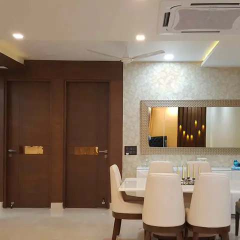Dining table with cream upholstered chairs beneath a large textured-frame mirror on floral wallpaper, flanked by twin dark-wood doors with brass inlay.