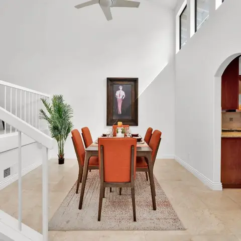 Double-height dining nook with a burnt-orange upholstered dining set beneath a staircase landing, opening to a kitchen through an arched doorway