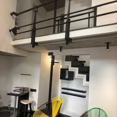 Double-height industrial loft with an exposed concrete mezzanine, black pipe railings, and yellow and green wire-frame accent chairs below the stair.