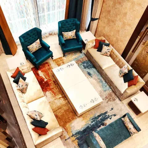 Double-height living room viewed from above, with cream tufted sofas and teal wingback chairs arranged around a bold abstract-pattern rug and a white
