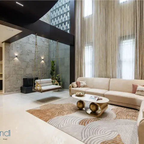 Double-height living room where a curved cream velvet sectional faces a brass-chain jhula hung against a carved leaf-relief stone wall