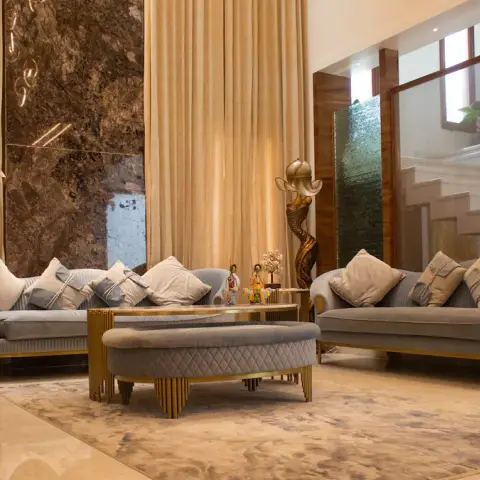 Double-height living room where two grey quilted-velvet sofas on fluted gold plinths face a book-matched brown marble slab panel between full-height beige
