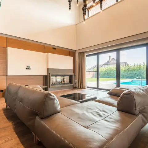Double-height living room with a leather sectional facing a built-in fireplace feature wall and floor-to-ceiling glass doors opening onto a pool deck.