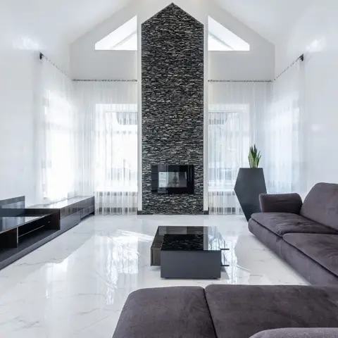 Double-height living room with a floor-to-ceiling stacked-stone fireplace wall beneath a vaulted skylight ceiling, sheer curtains