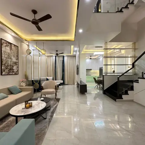 Double-height living room with a cream sofa and teal accent chair beneath gold cove lighting, opening to a dining nook