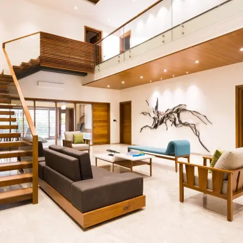 Double-height living room with a floating wood staircase, wood-clad mezzanine soffit, low wooden sofa set and a sculptural driftwood-style wall art.