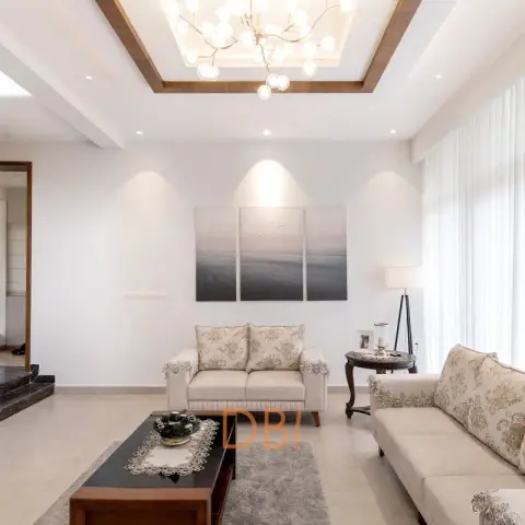 Double-height living room with a sculptural branch chandelier set into a wood-trimmed cove ceiling, floral lace-trimmed cream sofas, a black wood coffee table
