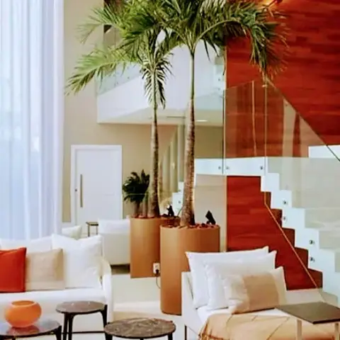 Double-height living room with a curved white sofa and orange accent pillow, tall potted palms flanking a floating staircase with glass balustrade and a rich