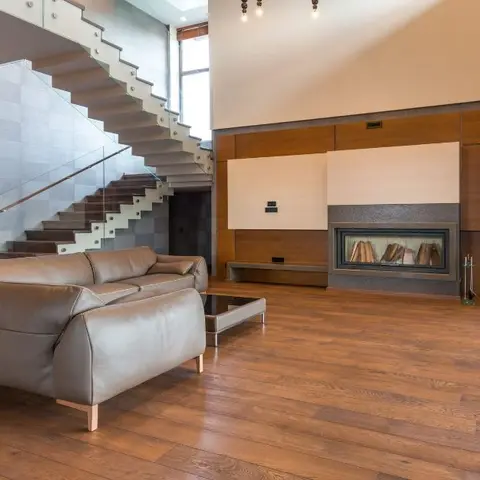 Double-height living room with a glass-railed staircase, a leather sectional sofa, and a built-in fireplace framed by wood wall panelling.