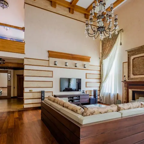 Double-height living room with a wood-beamed ceiling, crystal chandelier, and a raised marble fireplace flanked by mullioned wall panelling.