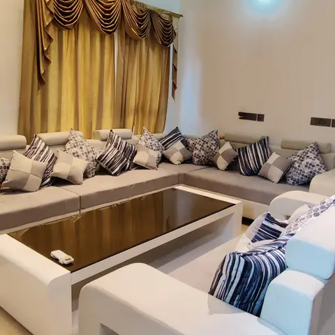 Double-height living room with a cascading chandelier, gold swag curtain valances, an L-shaped grey sectional sofa, and a glass-top white coffee table.