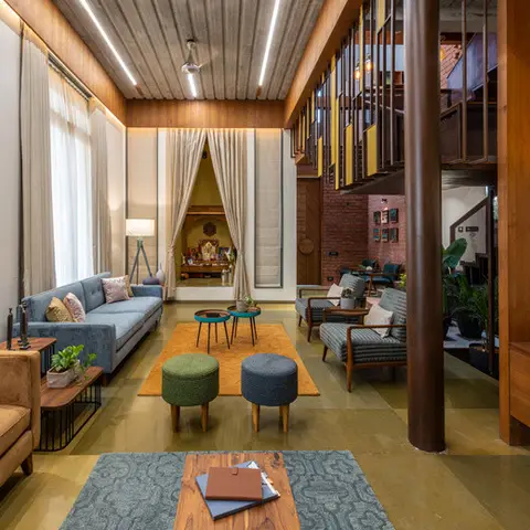 Double-height living room with an exposed wood-slat ceiling and mezzanine above, mismatched blue and mustard sofas, colorful poufs on a patterned rug