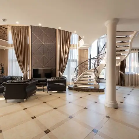 Double-height living room with black tufted leather sofas facing a quilted panel TV wall, a curved staircase and columns in the background.