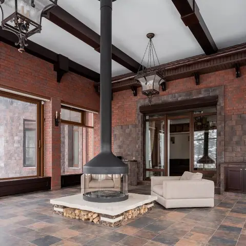 Double-height living room with exposed red brick walls, timber roof beams, a central black flue wood-burning stove on a raised stone hearth