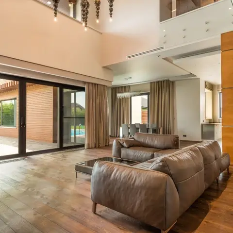 Double-height open-plan villa interior with a brown leather sofa in the foreground, a dining table and kitchen beyond, wide-plank wood flooring