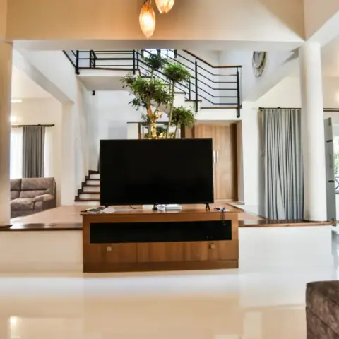 Double-height villa living hall shot low across a mirror-gloss cream tiled floor, with a teak TV console on a raised plinth and a tall indoor tree rising