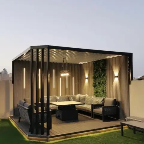Dusk view of a terrace pergola room: a black louvered aluminium frame lined on three sides with fluted wood cladding and vertical LED strips